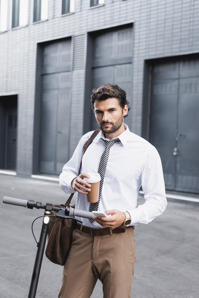 γενειοφόρος επιχειρηματίας με κοστούμι κρατώντας καφέ για να πάει και τη χρήση smartphone κοντά e-scooter  - Φωτογραφία, εικόνα