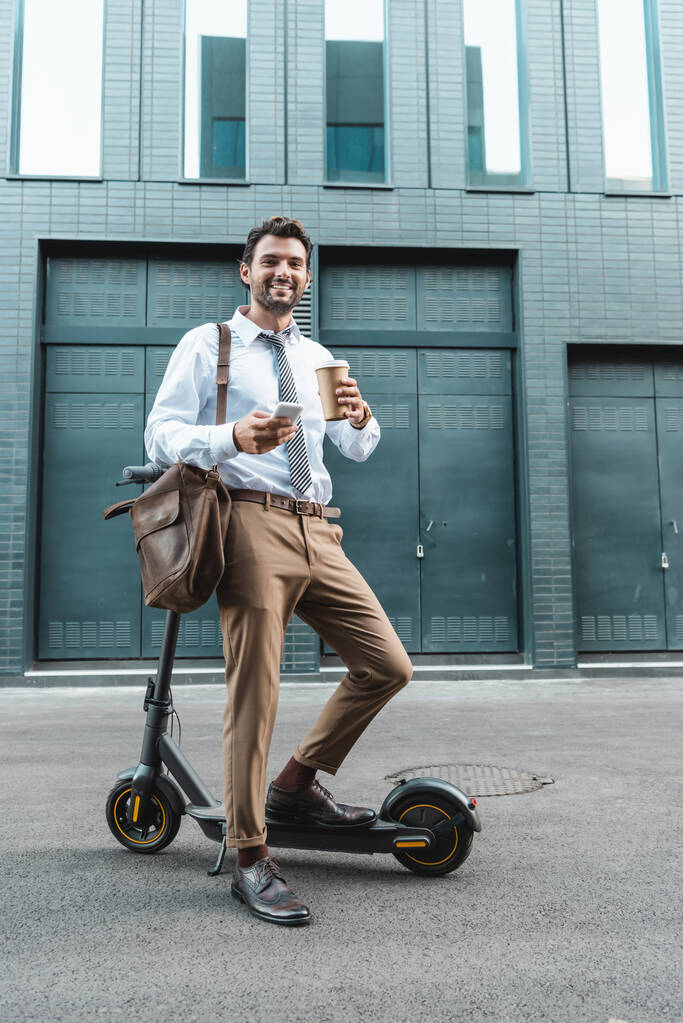piena lunghezza di uomo d'affari sorridente in abbigliamento formale in possesso di tazza di carta e sms su smartphone vicino e-scooter  - Foto, immagini