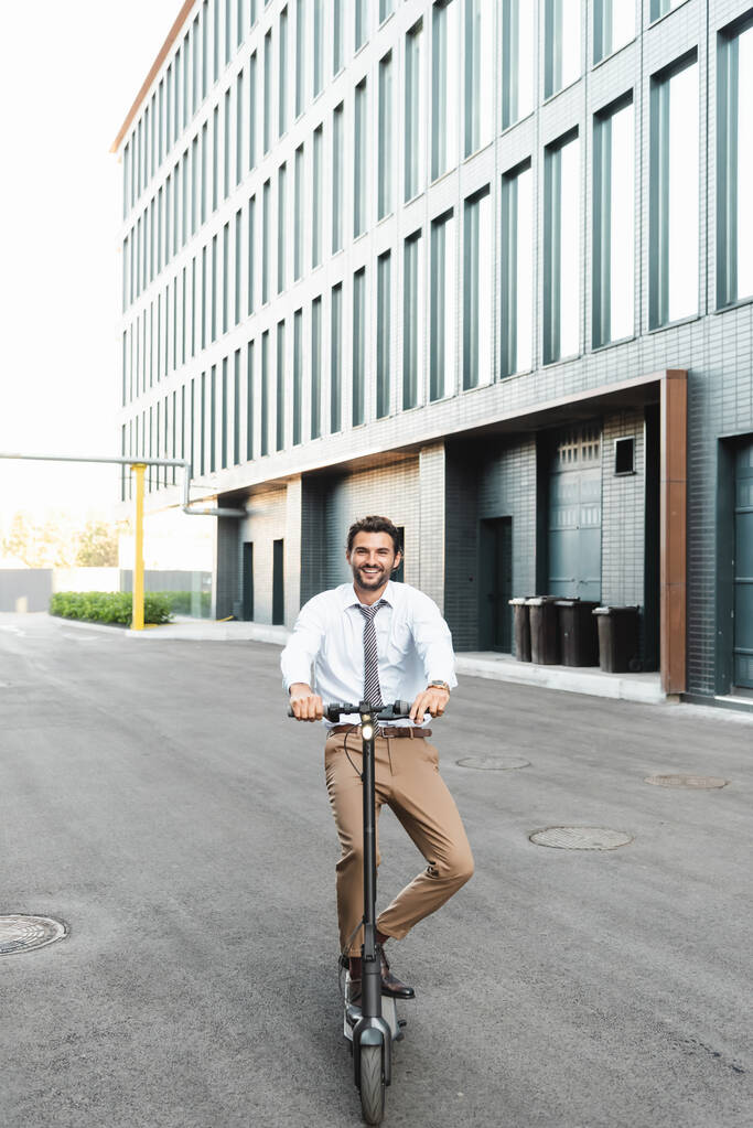 Счастливый бизнесмен в полный рост катается на электроскутере возле здания  - Фото, изображение