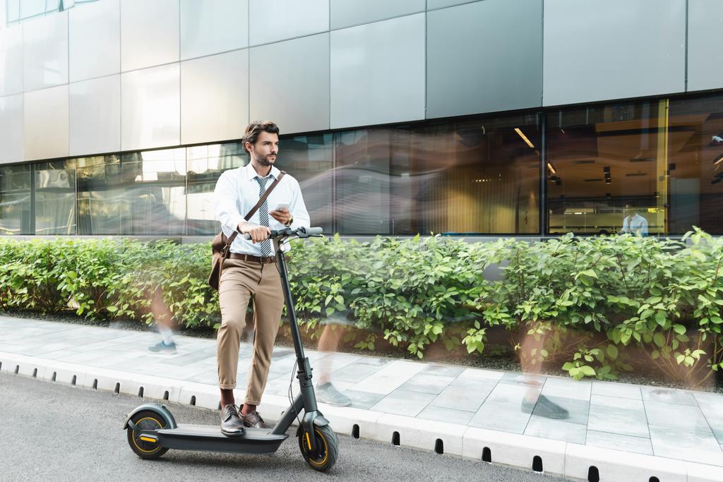 pohybové rozmazání podnikatel drží smartphone, zatímco stojí v blízkosti e-scooter, rostliny a budovy  - Fotografie, Obrázek