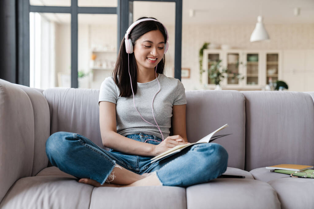 Imagen de una mujer asiática feliz usando auriculares y anotando notas mientras está sentada en el sofá en casa - Foto, Imagen