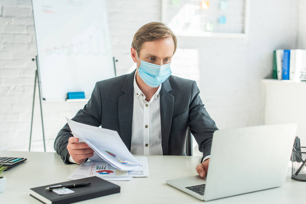 Empresario en máscara médica, sosteniendo papeles y usando computadora portátil, mientras está sentado en el lugar de trabajo en un fondo borroso - Foto, imagen