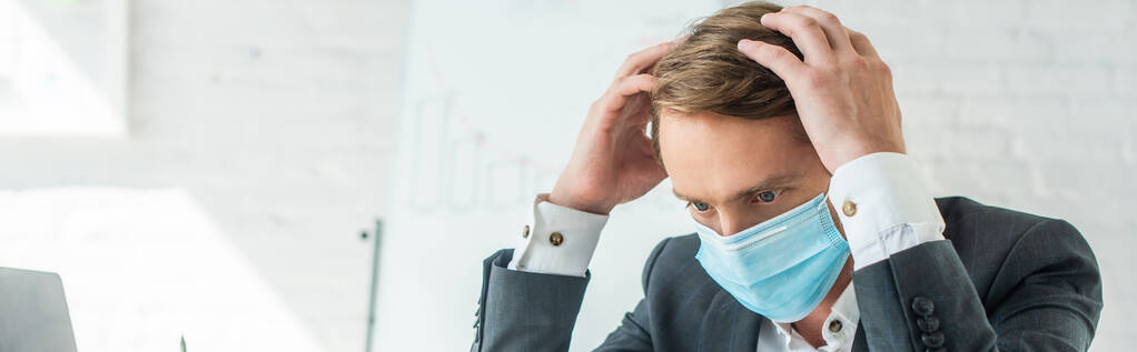 Biznesmen w masce medycznej trzymający się za ręce przy głowie z rozmazanym flipchartem na tle, baner - Zdjęcie, obraz