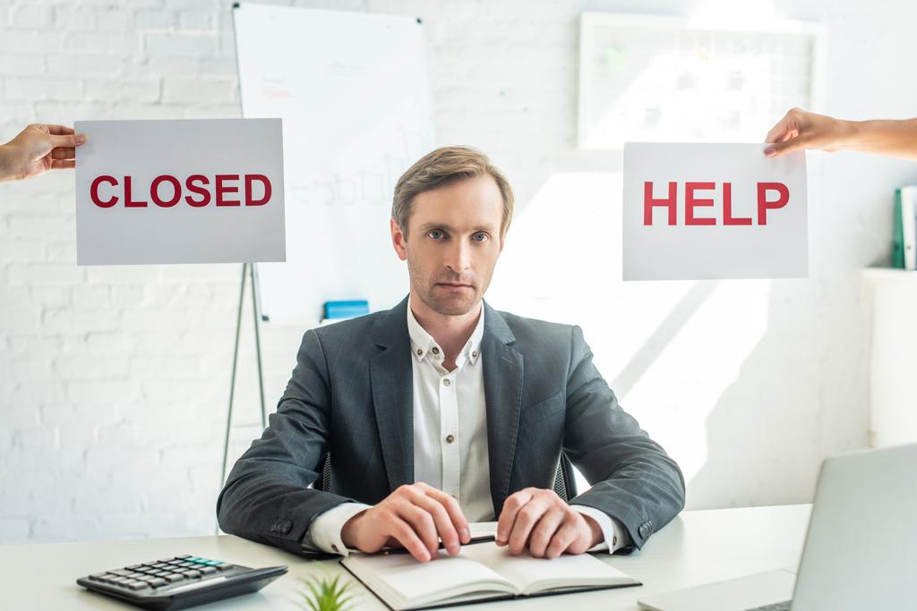 Businessman looking at camera, while sitting at workplace near signs with closed and help lettering on blurred background - Photo, Image