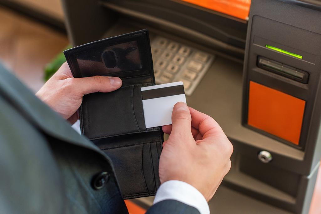Обрізаний вид бізнесмена, що кладе кредитну картку в гаманець, стоячи поруч з атмом на розмитому передньому плані
 - Фото, зображення