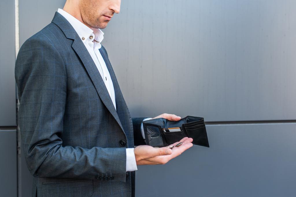 Vista recortada del hombre de negocios con monedas en la palma, sosteniendo la cartera vacía sobre fondo texturizado gris - Foto, imagen