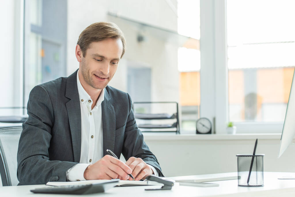 Χαμογελαστός επιχειρηματίας που γράφει στο σημειωματάριο, ενώ κάθεται στο τραπέζι με θολή παράθυρο στο παρασκήνιο - Φωτογραφία, εικόνα