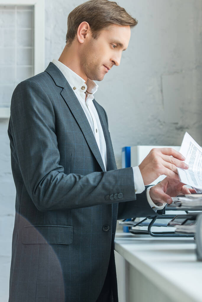 Boční pohled zaostřeného podnikatele, jak se dívá do papírů, zatímco stojí u podnosu s dokumenty na parapetu - Fotografie, Obrázek