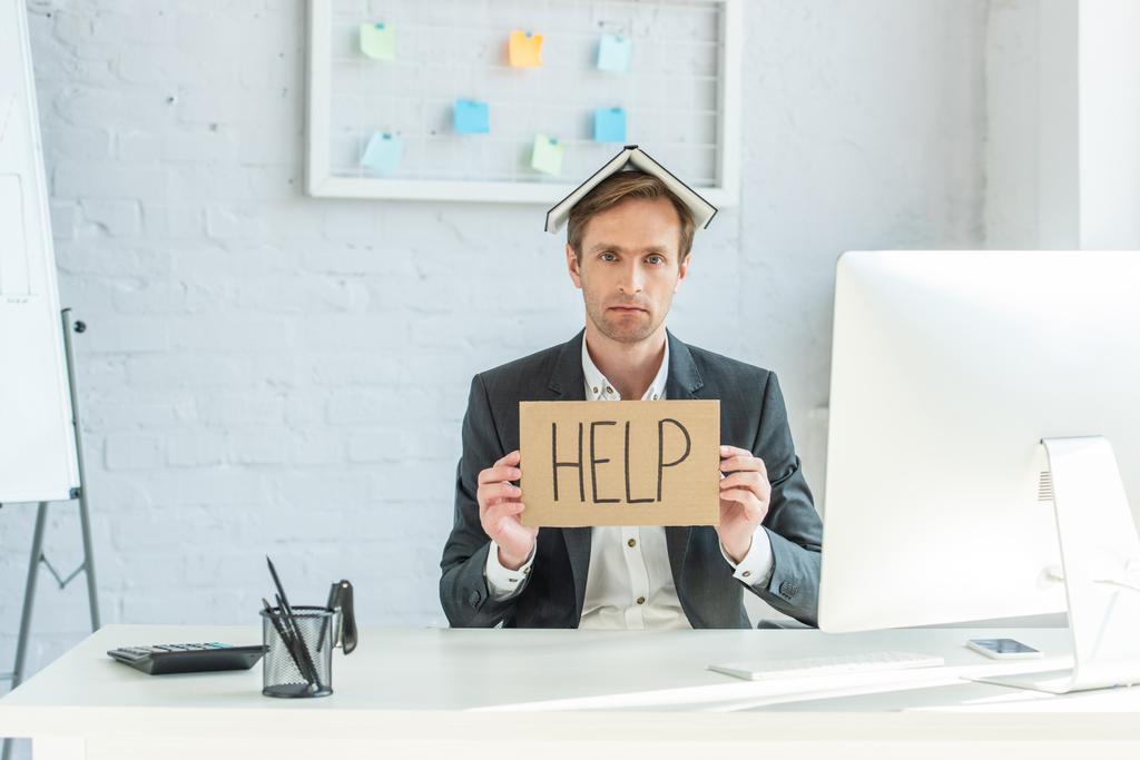 Sad businessman with notebook on head, holding cardboard with help lettering, while sitting at workplace - Photo, Image