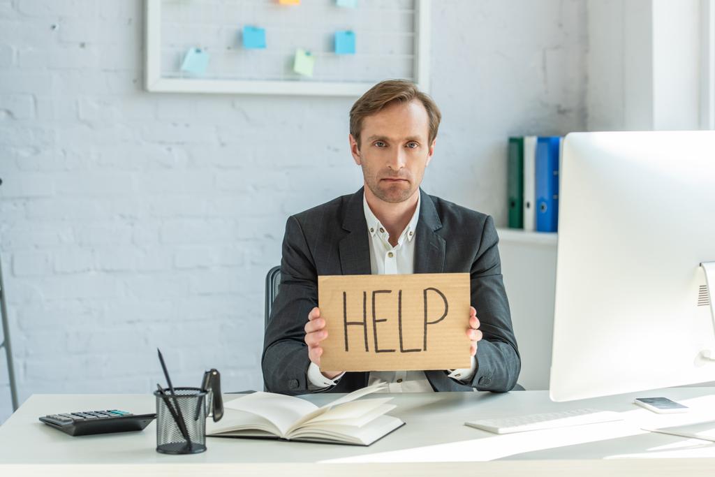 Sad businessman looking at camera and holding cardboard with help lettering, while sitting at workplace - Photo, Image