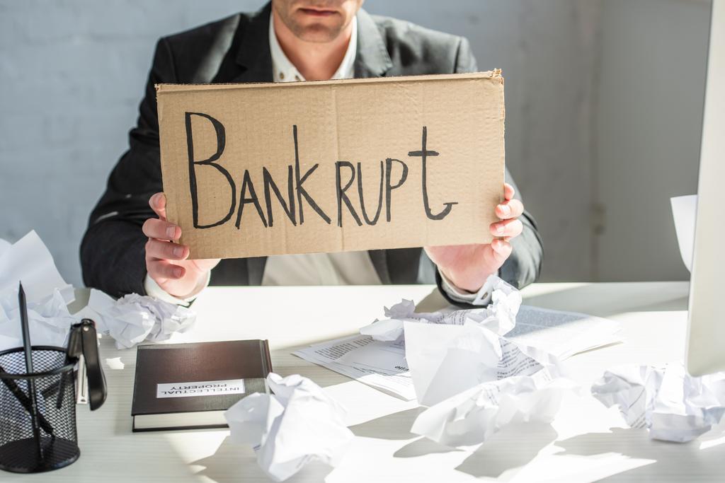 Cropped view of businessman holding cardboard with bankrupt lettering, while sitting at desk with crumpled papers - Photo, Image
