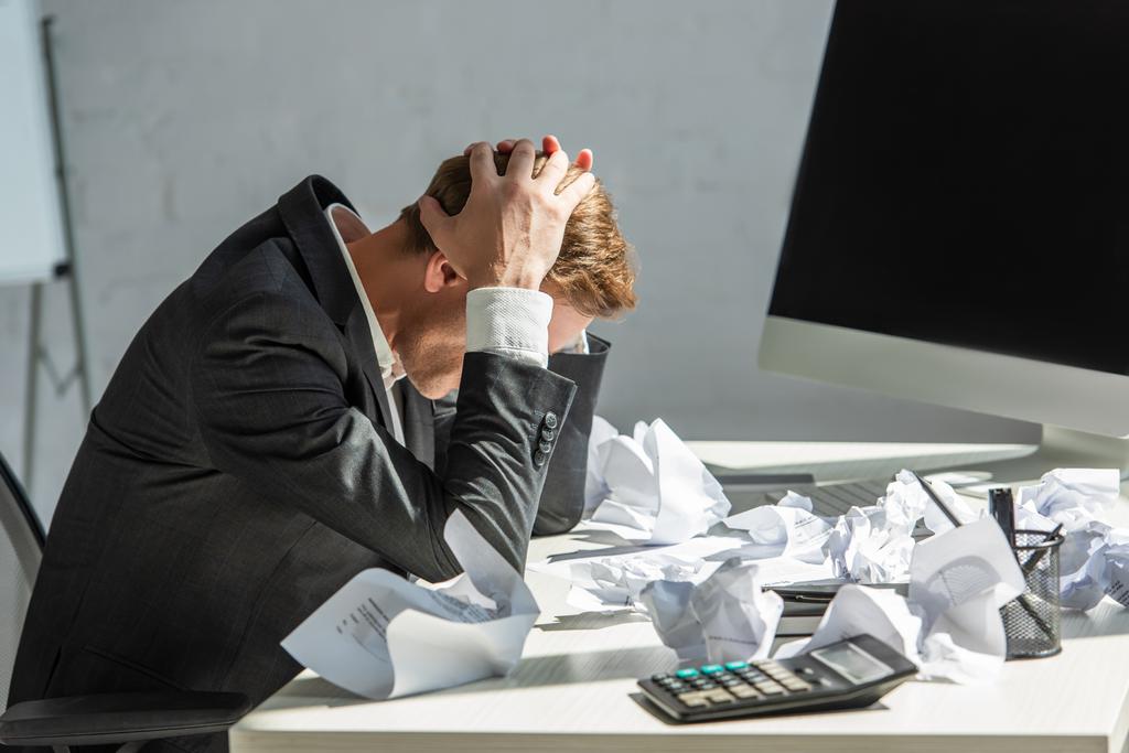 Gestresster Geschäftsmann mit den Händen auf dem Kopf, am Arbeitsplatz sitzend mit zerbröckelten Papieren auf verschwommenem Hintergrund - Foto, Bild