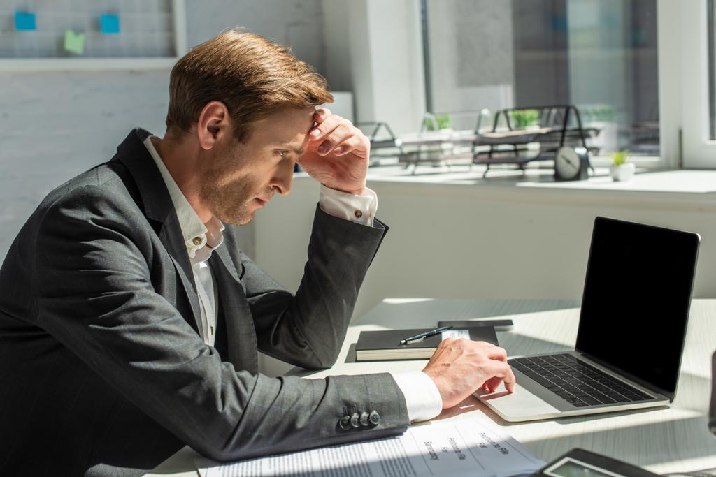 Przygnębiony biznesmen z ręką na czole, korzystający z laptopa, siedzący w miejscu pracy z rozmazanym biurem na tle - Zdjęcie, obraz