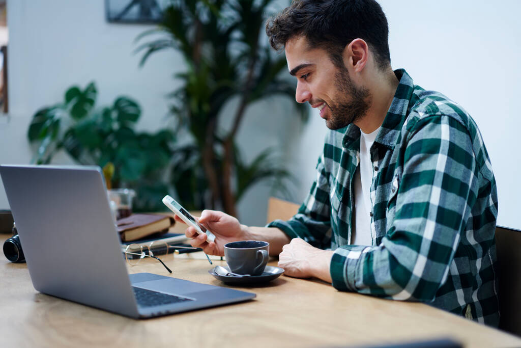 Zijaanzicht van positieve bebaarde etnische mannelijke freelancer in casual outfit en brillen browsen smartphone tijdens het werken op afstand op laptop in cafe - Foto, afbeelding