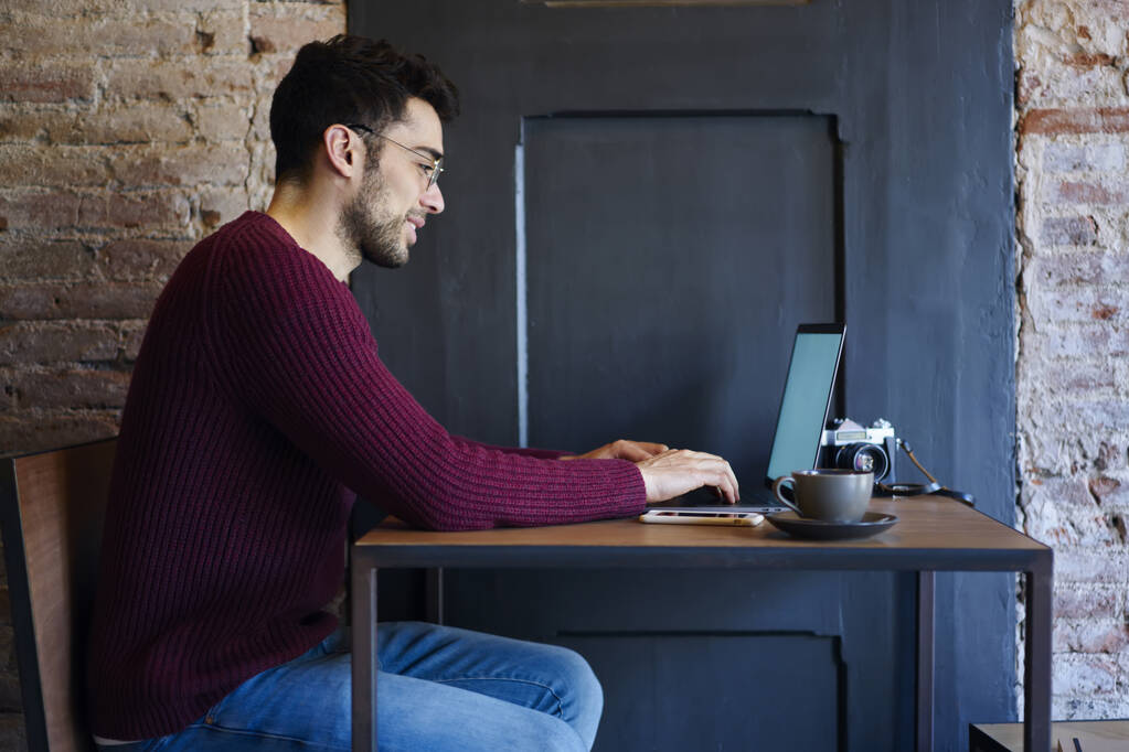 Boční pohled na pozitivní mladý vousatý muž fotograf v purpurovém svetru a džíny pracující na notebooku z jasné útulné kavárny - Fotografie, Obrázek