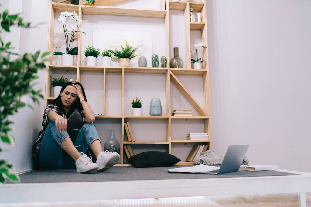 Full length of female worker in casual outfit sitting on floor with laptop and documents while experiencing headache and leaning on hand - Photo, Image