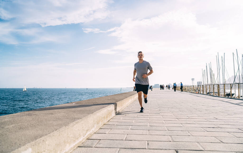 Caucasian male runner in casual sportswear enjoying morning cardio workout for keeping healthy lifestyle, determined man with perfect body shape jogging at sea embankment during sportive activity - Photo, Image