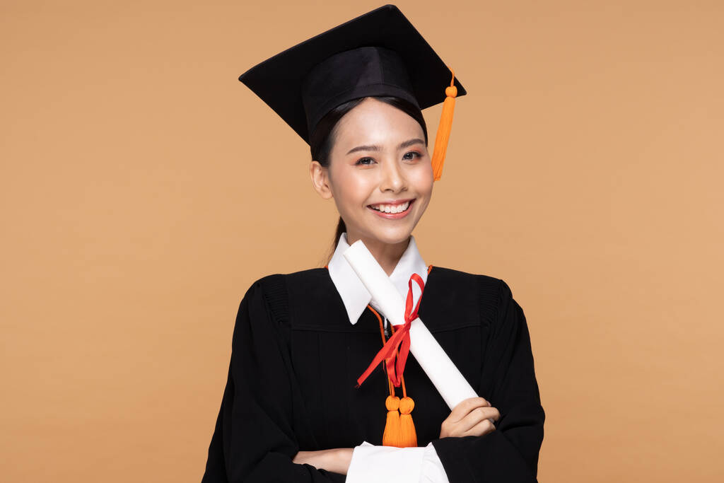 Heureuse diplômée asiatique femme en bonnet et gowm tenant Certificat ou diplôme sur fond beige, Graduation Concept - Photo, image