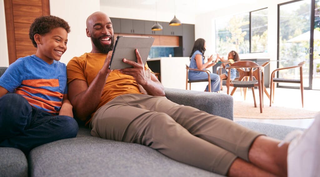 Familia afroamericana en casa con padre e hijo sentados en sofá en casa usando tableta digital - Foto, imagen
