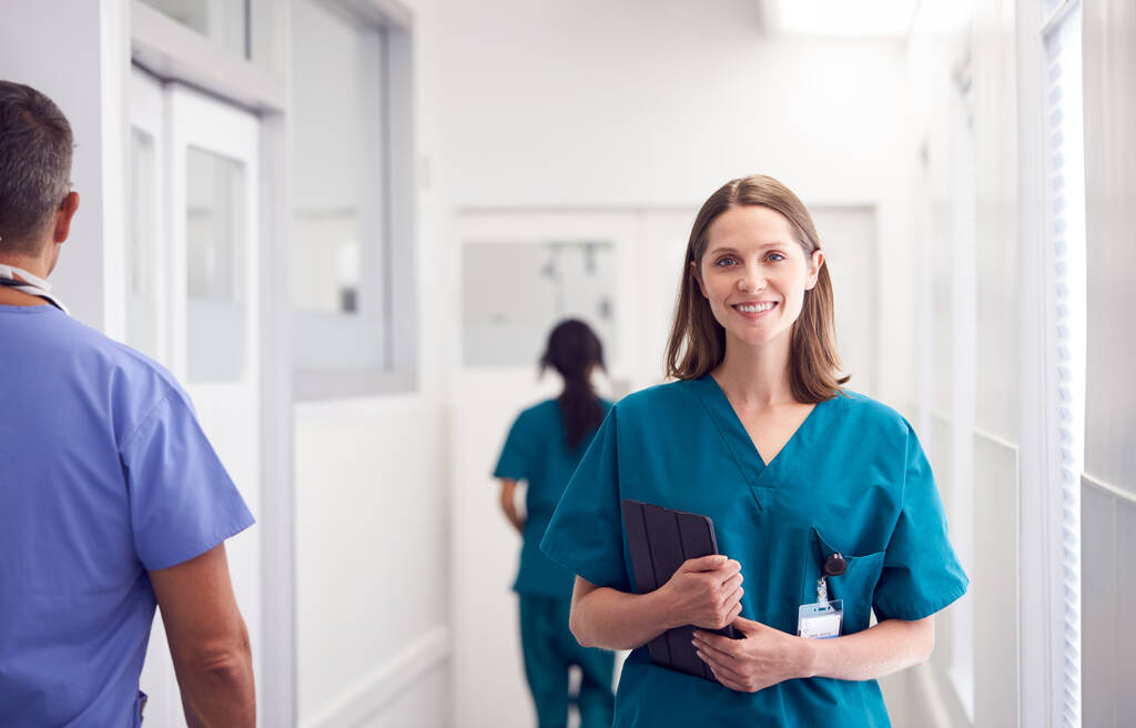 Ritratto di sorridente medico donna che indossa scrub nel corridoio ospedaliero occupato con tablet digitale - Foto, immagini