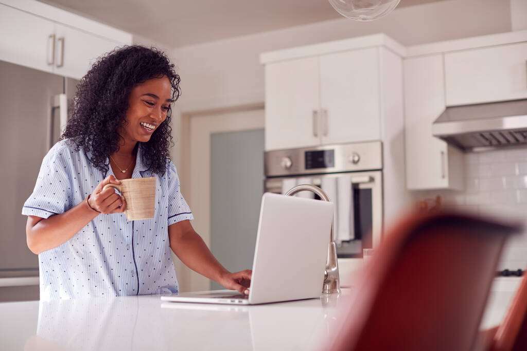 Woman Wearing Pyjamas Standing In Kitchen Working From Home On Laptop - Photo, Image