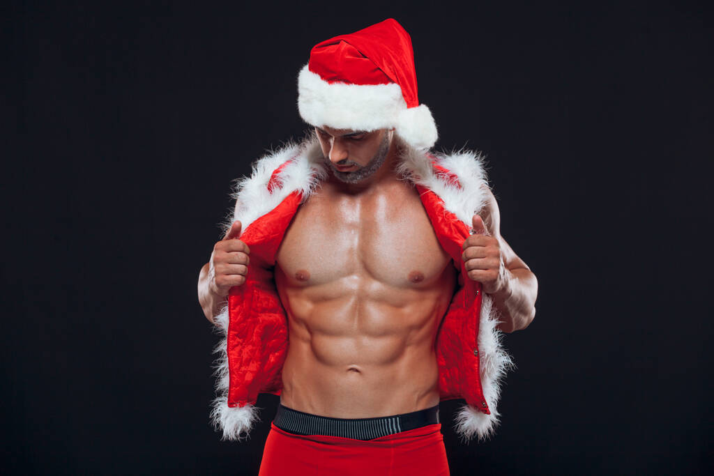 Seksi Noel Baba. Vücut geliştirici genç yakışıklı Noel Baba kılıklı gülümseme Noel Baba şapkası takıyor yeni yıl arifesinde karın kaslarını gösteriyor ve Noel tatilinin kara arka planında. - Fotoğraf, Görsel