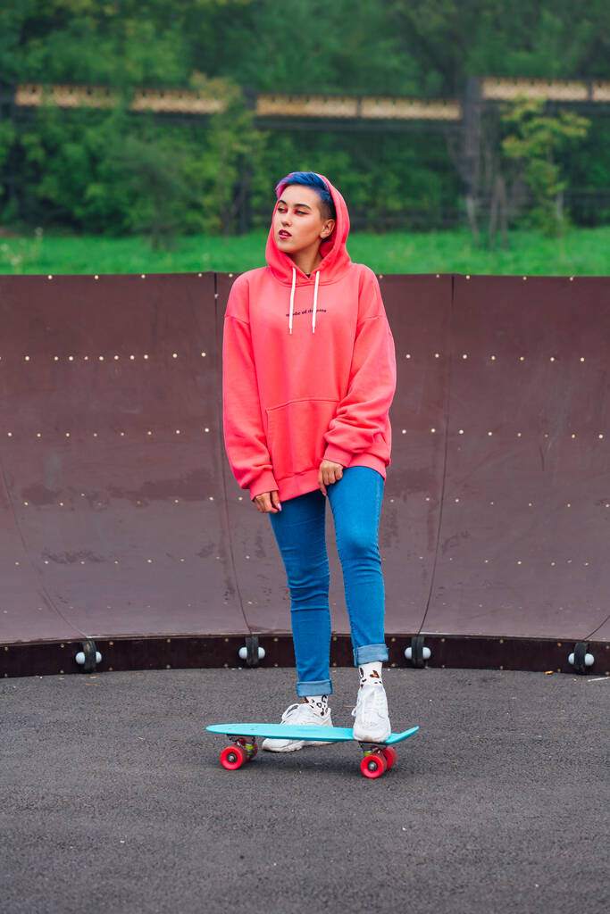 Porträt eines trendigen hübschen jungen Mädchens in rosa Kapuzenpulli und Jeans, das mit seinem blauen Plastik-Skateboard neben dem Skateboard-Platz steht. - Foto, Bild