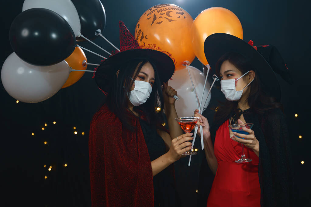 portrét dvou asijských žen v kostýmu čarodějnic s ochrannou obličejovou maskou na ochranu koronaviru covid-19 se sklenicemi koktejlového nápoje a balónem na halloween party - Fotografie, Obrázek
