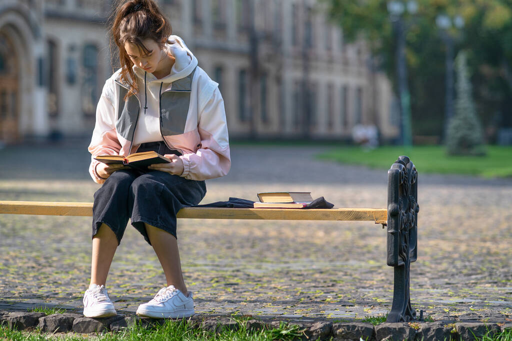 Adolescente joven de moda sentada estudiando en un banco en un parque leyendo un libro con copyspace y fondo de edificio histórico - Foto, imagen