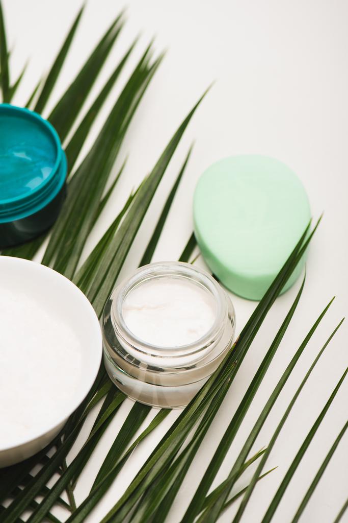 recipiente con crema cosmética casera, exfoliante, tazón y jabón cerca de hojas de palma en blanco - Foto, Imagen