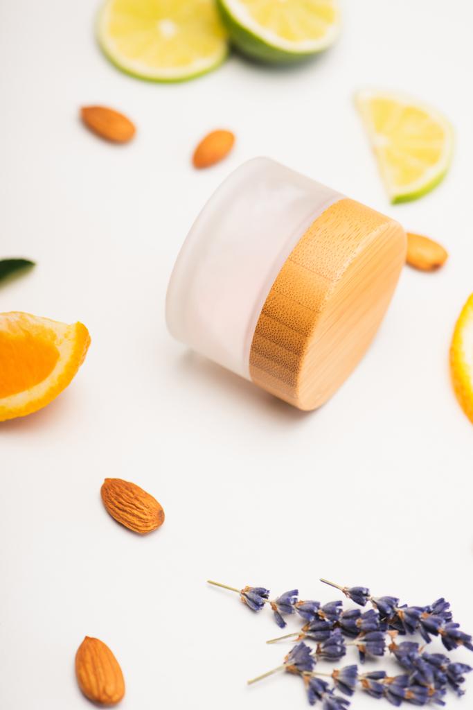 δοχείο σπιτικής καλλυντικής κρέμας κοντά σε φέτες πορτοκάλι, λάιμ και λεμόνι, αμύγδαλα και κλαδιά λεβάντας σε λευκό θολή φόντο - Φωτογραφία, εικόνα
