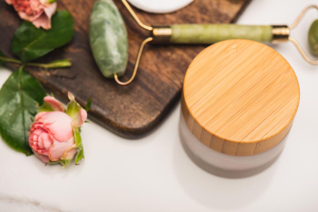 container van cosmetische crème in de buurt van snijplank met jade roller, en thee rozen op wazig witte achtergrond  - Foto, afbeelding