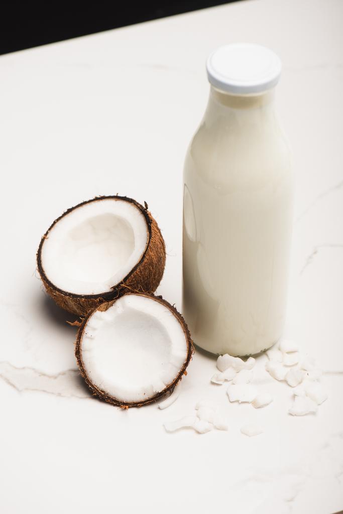butelka mleka kokosowego w pobliżu połówek i płatków na białej powierzchni wyizolowana na czarno - Zdjęcie, obraz