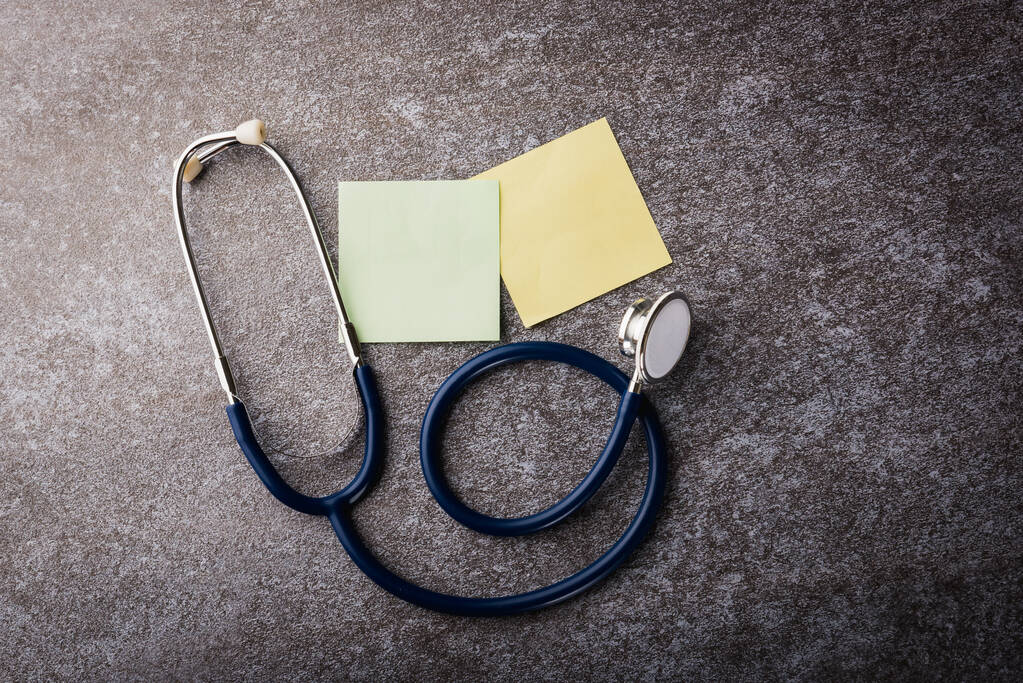 Yläpuolella lääkäri toimisto pöytä pöytä tyhjä paperi tikku huomata keltainen ja vihreä ja stetoskooppi betonitaustalla, Muistutus käsite - Valokuva, kuva