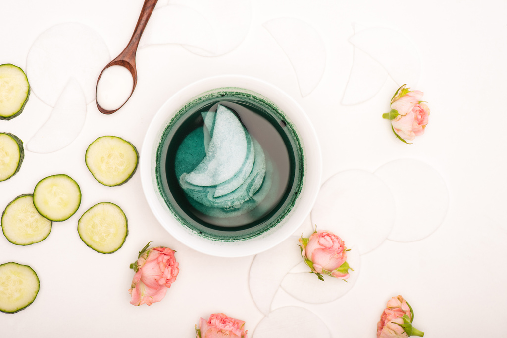 vue du dessus du bol avec tampons toniques et coton, roses de thé, tranches de concombre frais et cuillère à crème cosmétique sur blanc - Photo, image