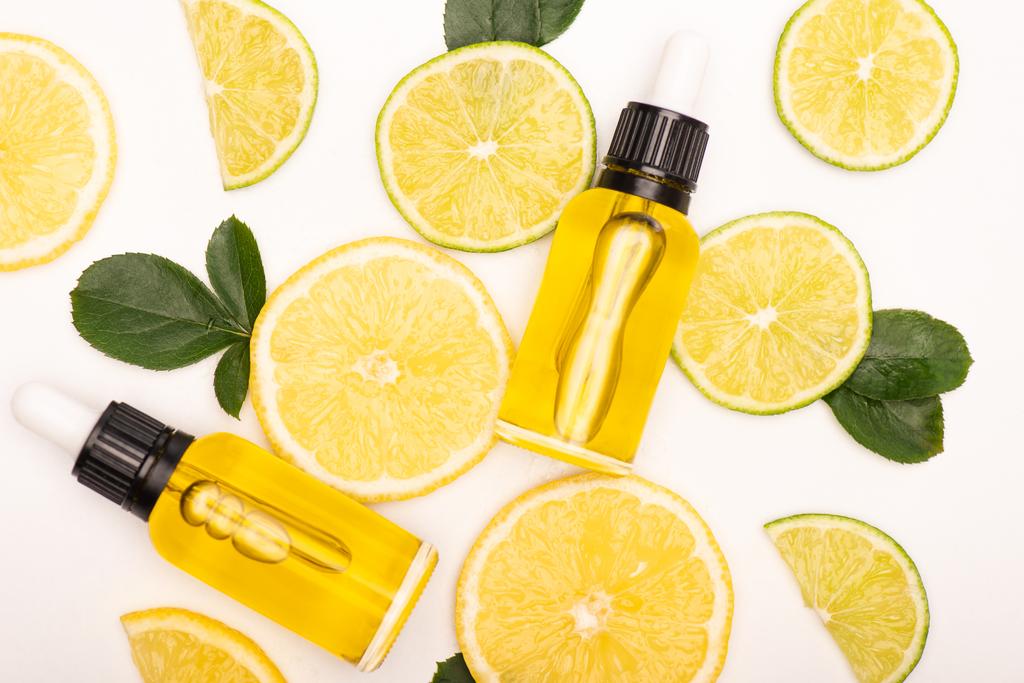 vrchní pohled na láhve s citrusovým esenciálním olejem v blízkosti citronu a citrusových plátků v blízkosti růžových lístků na bílém - Fotografie, Obrázek