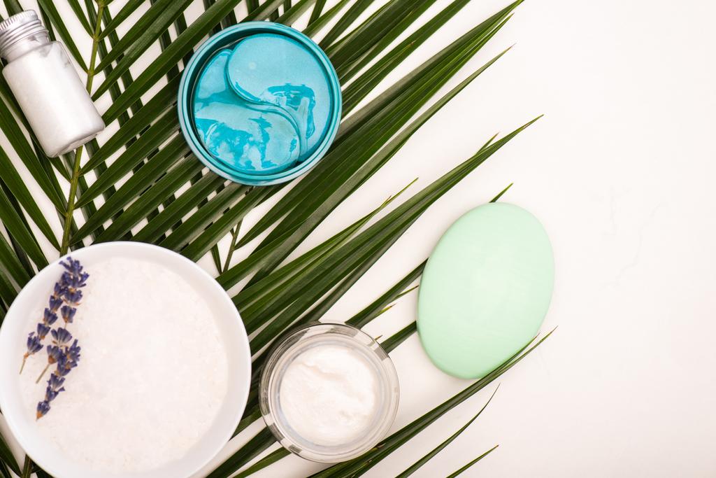 vue du dessus du bol avec lavande, crème cosmétique, gommage et savon près des feuilles de palmier sur blanc - Photo, image