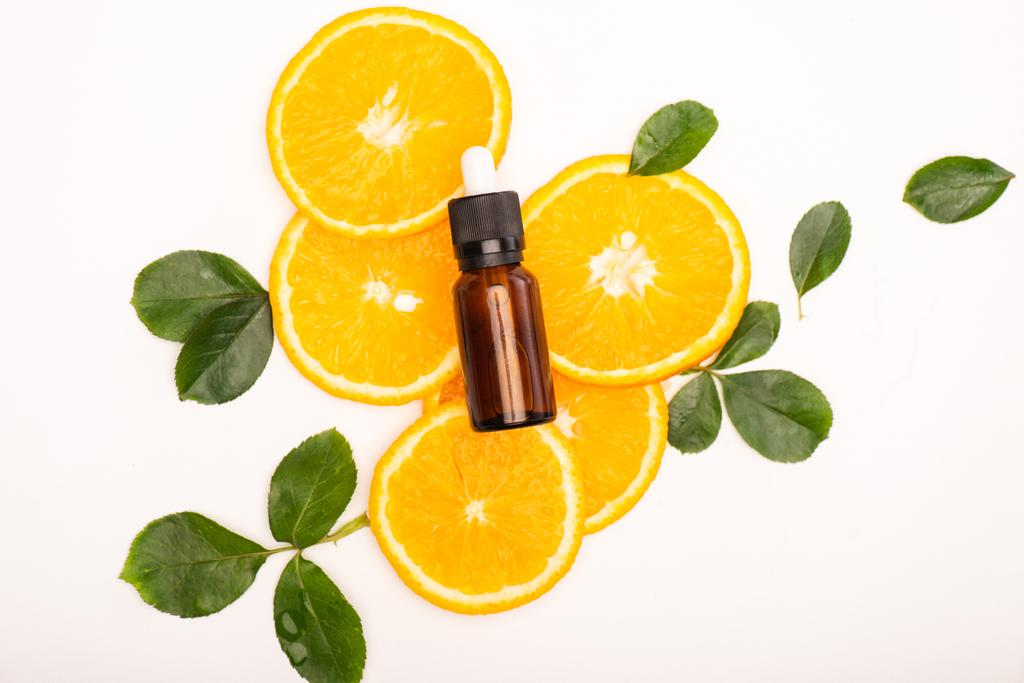 bovenaanzicht van fles met citrus essentiële olie en sappige sinaasappelschijfjes in de buurt van rozenblaadjes op wit - Foto, afbeelding