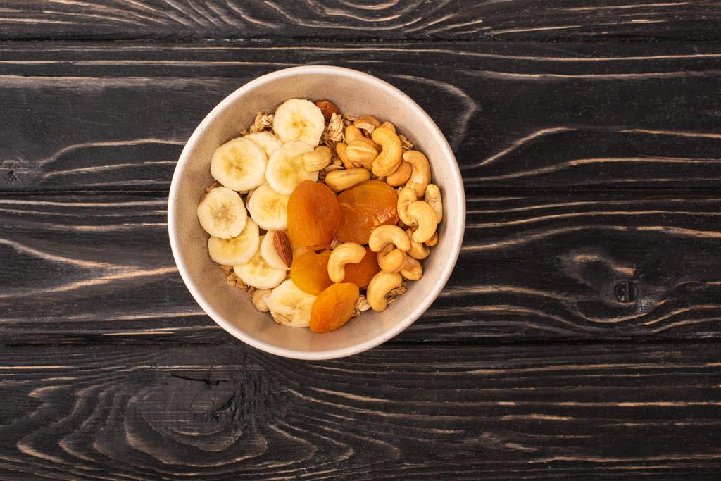 horní pohled na lahodnou granolu s ořechy, banány a sušenými meruňky na dřevěném černém povrchu - Fotografie, Obrázek