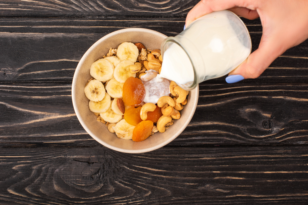 rajattu näkymä nainen lisäämällä jogurtti herkullinen mysli pähkinät, banaani ja kuivatut aprikoosit puinen musta pinta - Valokuva, kuva