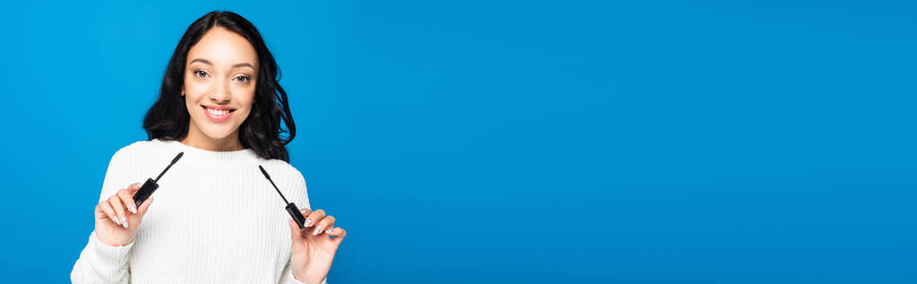 улыбающаяся брюнетка женщина держит тушь и кисть изолированы на синий, баннер - Фото, изображение