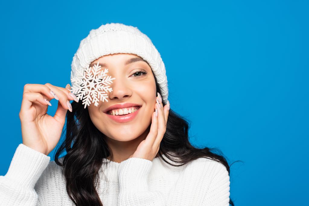 cheerful woman in knitted hat holding decorative snowflake near eye isolated on blue - Photo, Image