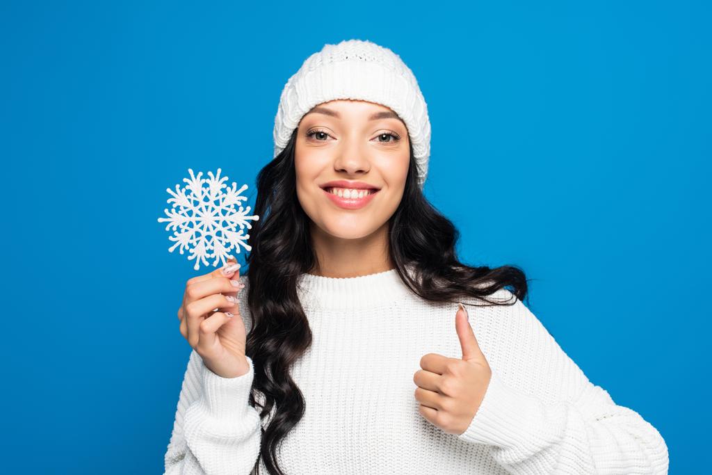 mujer feliz en sombrero de punto con copo de nieve decorativo y mostrando el pulgar hacia arriba aislado en azul - Foto, Imagen