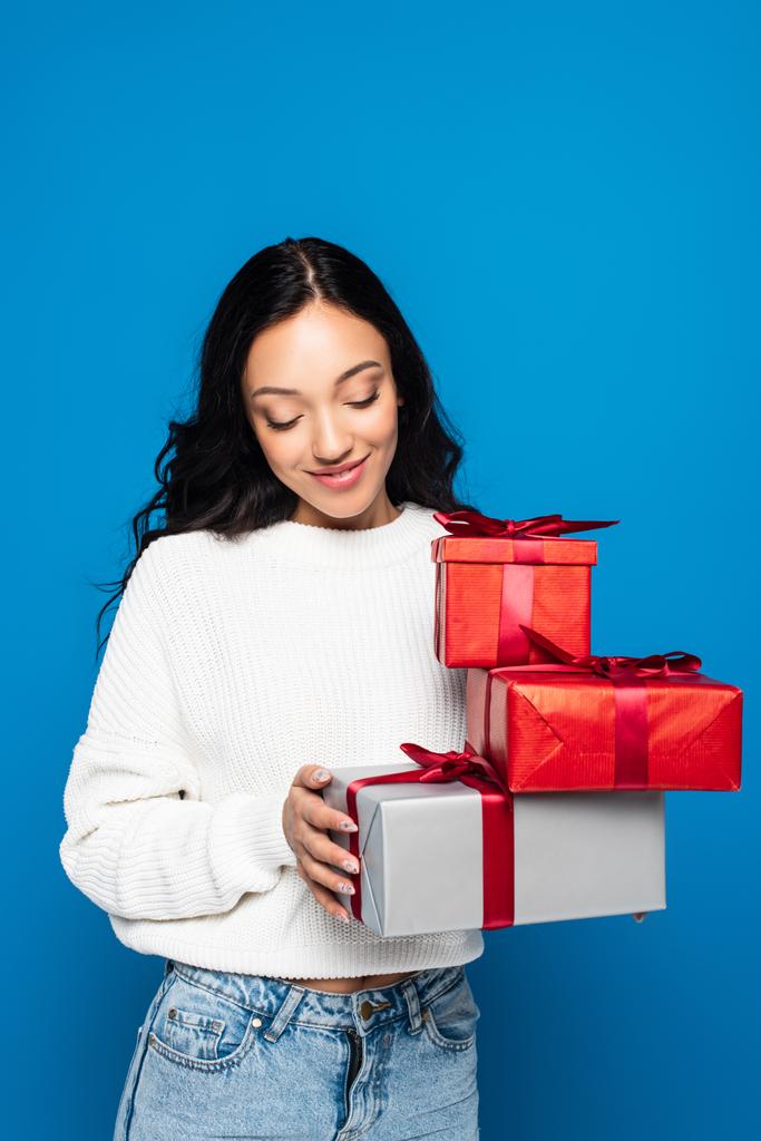 щаслива молода жінка в трикотажному светрі тримає подарункові коробки ізольовані на синьому
 - Фото, зображення