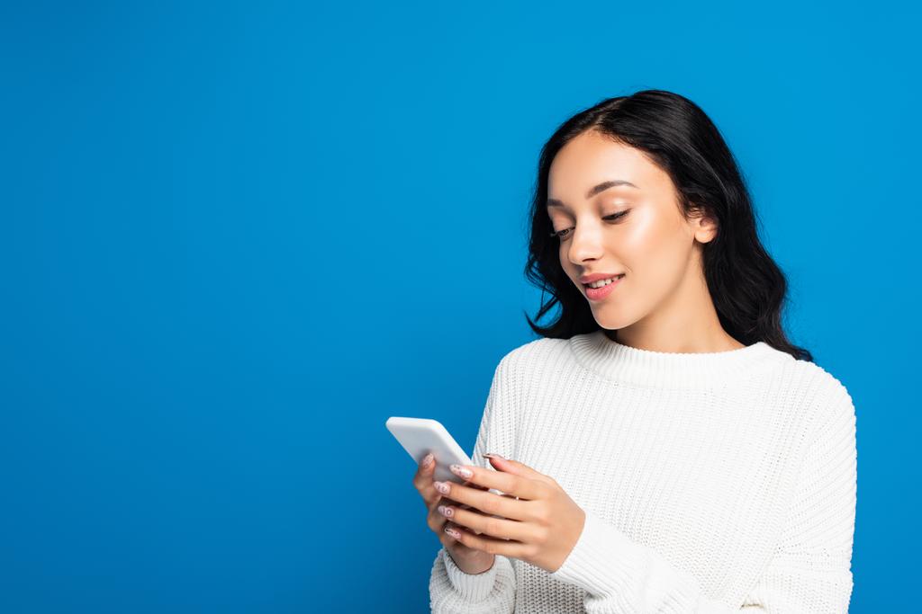 young woman in warm sweater using smartphone isolated on blue - Photo, Image