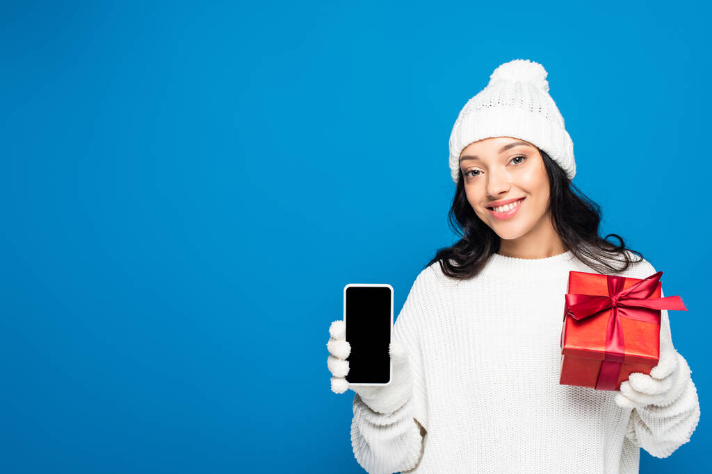 щаслива жінка в трикотажному капелюсі і рукавичках тримає подарункову коробку і смартфон з порожнім екраном ізольовано на синьому
 - Фото, зображення