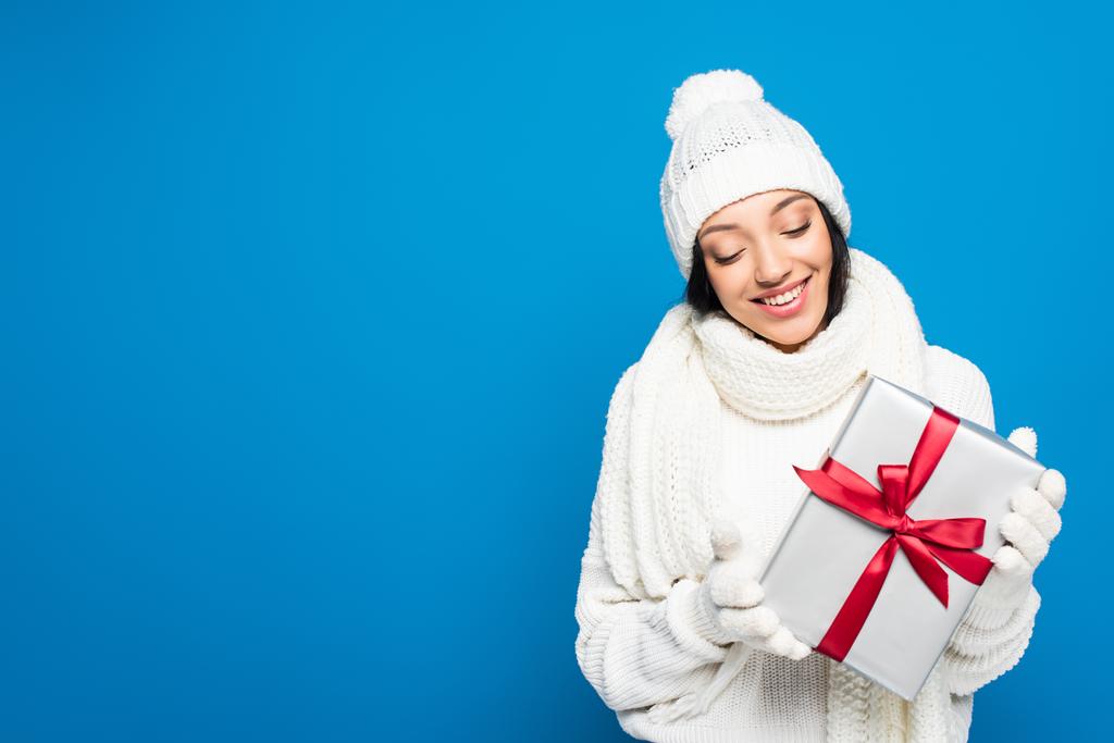 femme heureuse en tenue d'hiver tenant cadeau de Noël isolé sur bleu - Photo, image