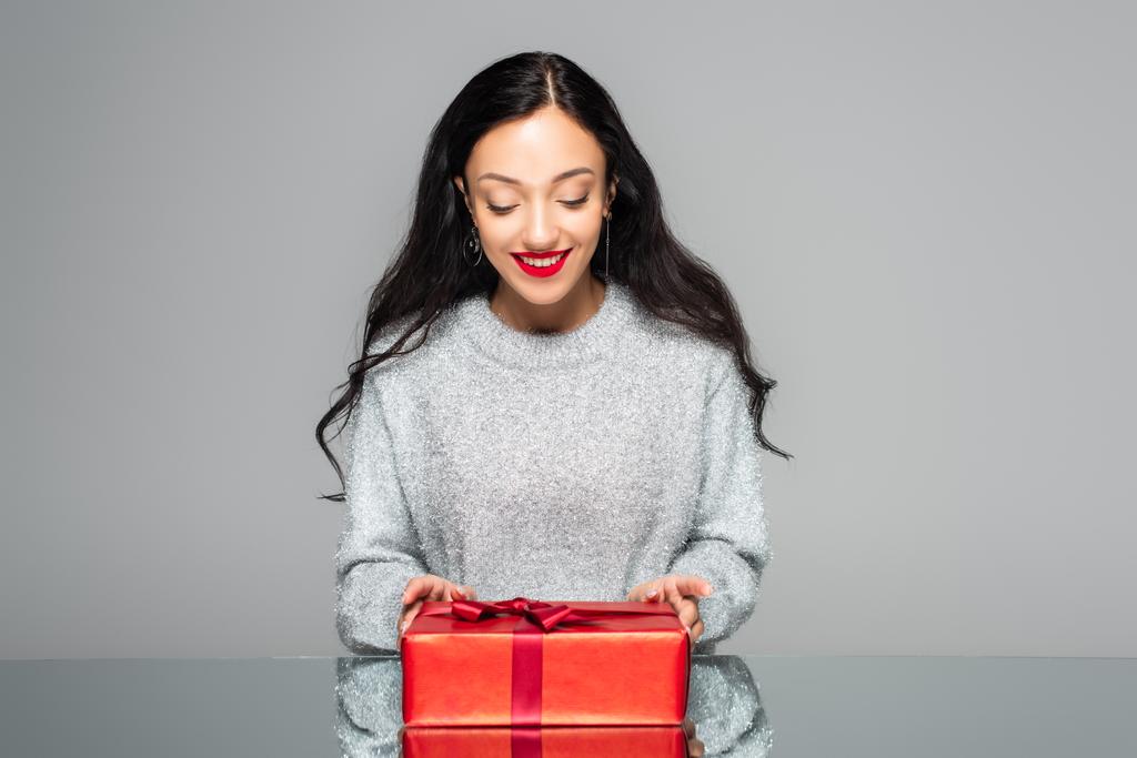 usmívající se žena s červenými rty při pohledu na dárkové krabici izolované na šedé - Fotografie, Obrázek
