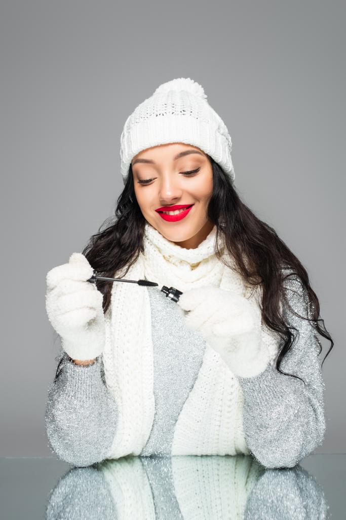 joyful and brunette woman in winter outfit holding mascara isolated on grey - Photo, Image