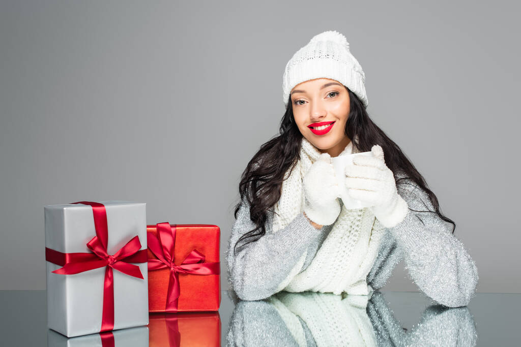 fröhliche und brünette Frau im Winter-Outfit mit Tasse isoliert auf grau - Foto, Bild